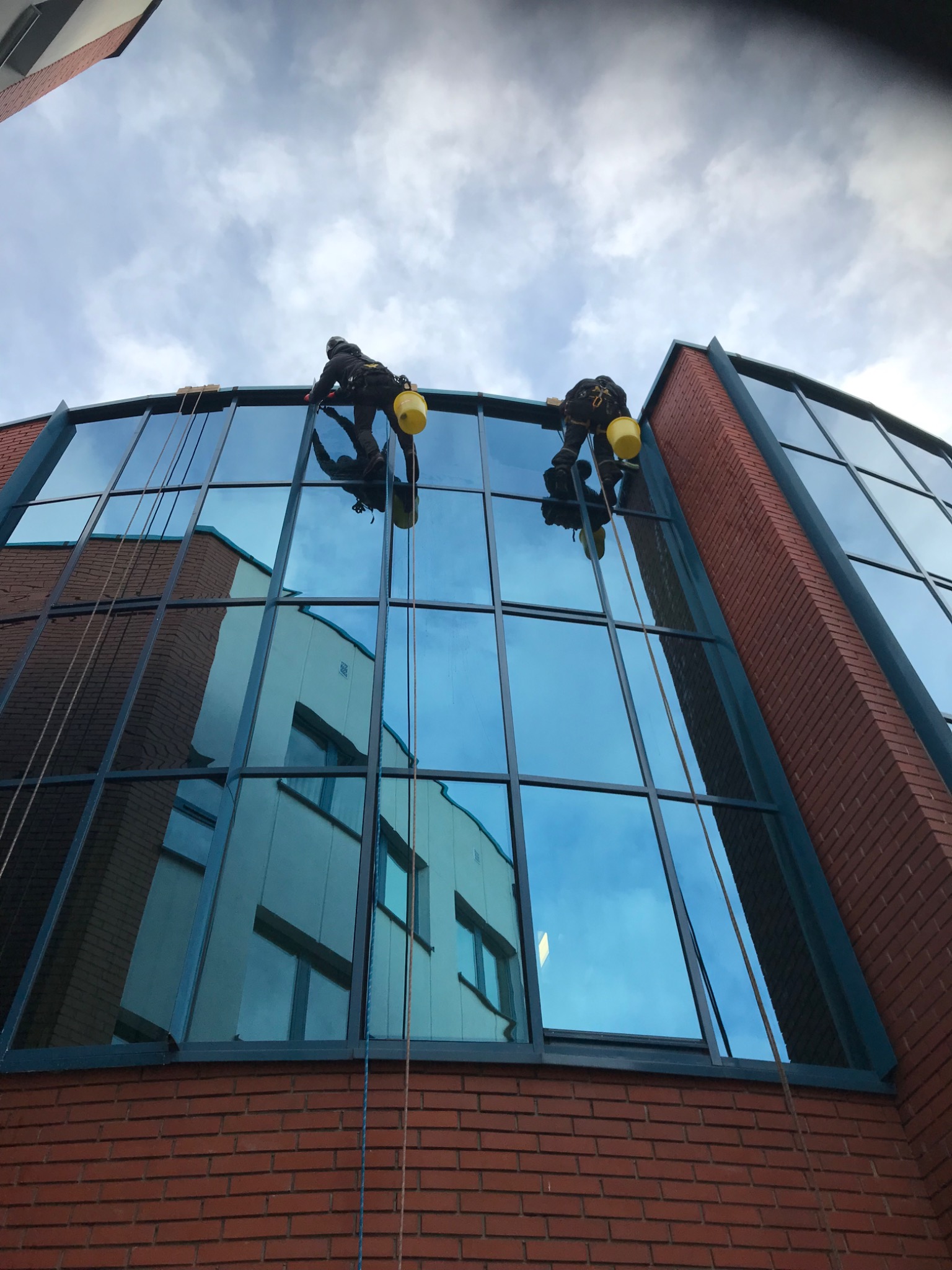Dwóch alpinistów myje okna na zakrzywionej fasadzie biurowca z czerwonej cegły, widoczne odbicie budynku naprzeciwko.