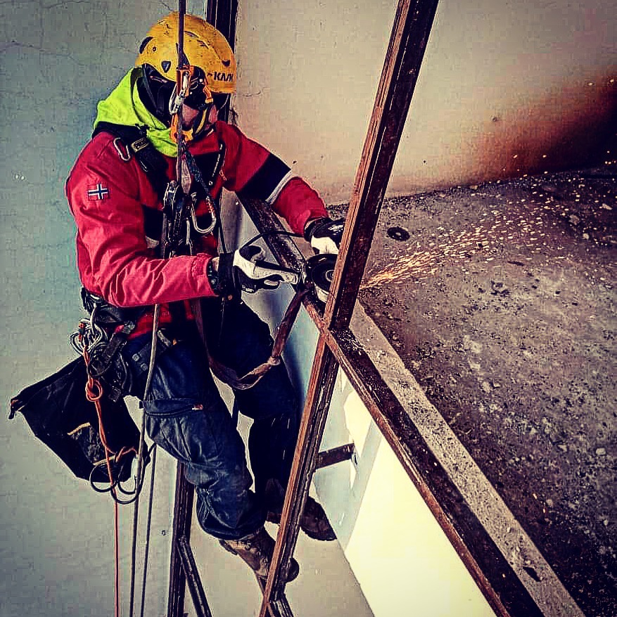 Pracownik w kasku i uprzęży, z flagą Norwegii na ramieniu, szlifuje metalową konstrukcję na wysokości, iskry lecą w tle brudnej ściany.