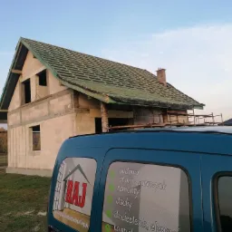 Budowa domu w stanie surowym otwartym z widocznym dachem w trakcie układania, na pierwszym planie samoch&oacute;d firmy budowlanej z reklamą usług.