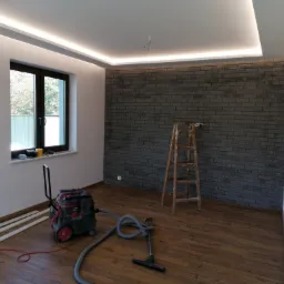 Pokój po remoncie z drewnianą podłogą, ścianą z szarej cegły, oświetleniem LED na suficie, drabina drewniana oparta o ścianę, odkurzacz przemysłowy i listwy przypodłogowe przy ścianie.
