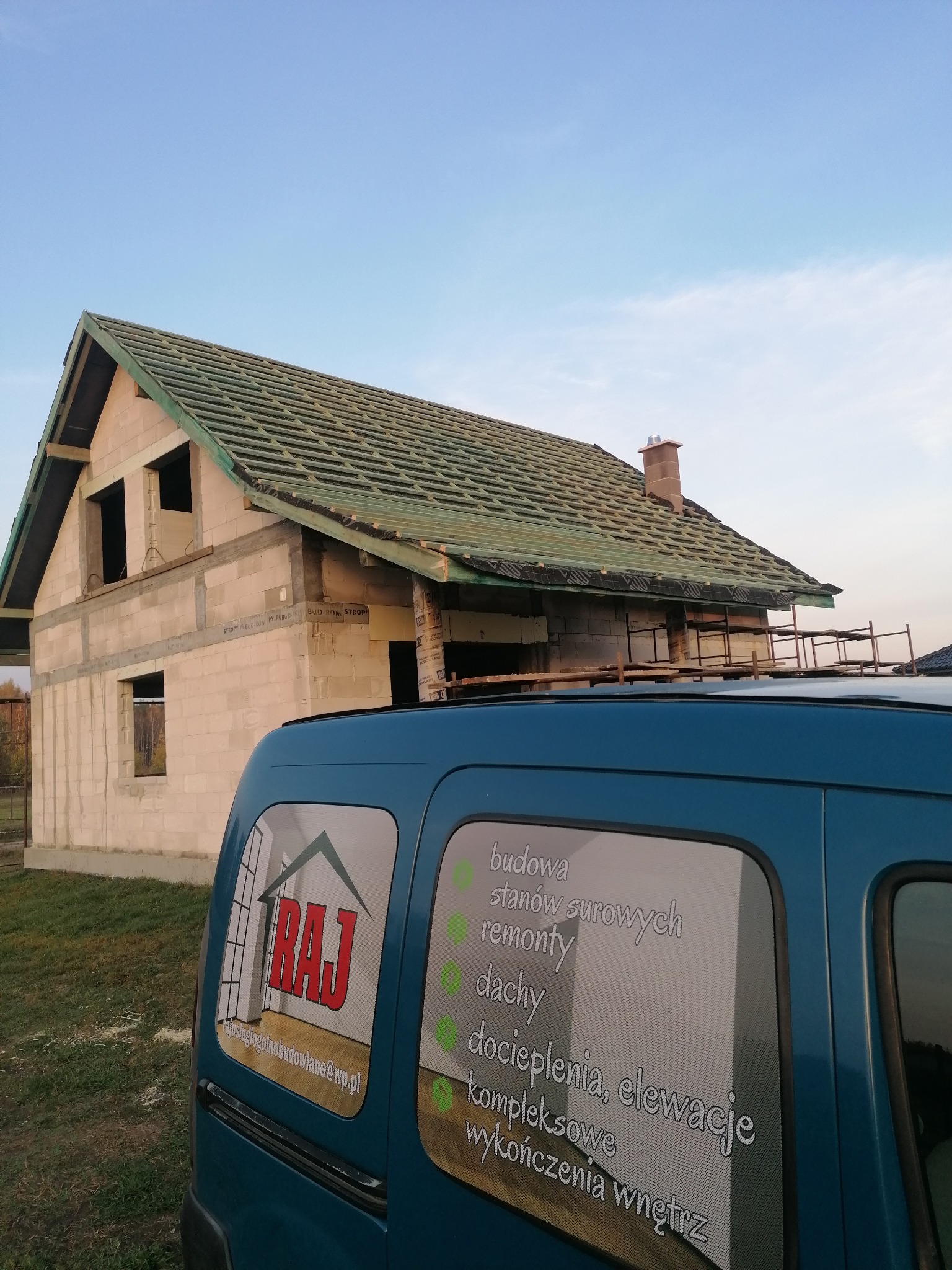 Budowa domu w stanie surowym otwartym z widocznym dachem w trakcie układania, na pierwszym planie samochód firmy budowlanej z reklamą usług.