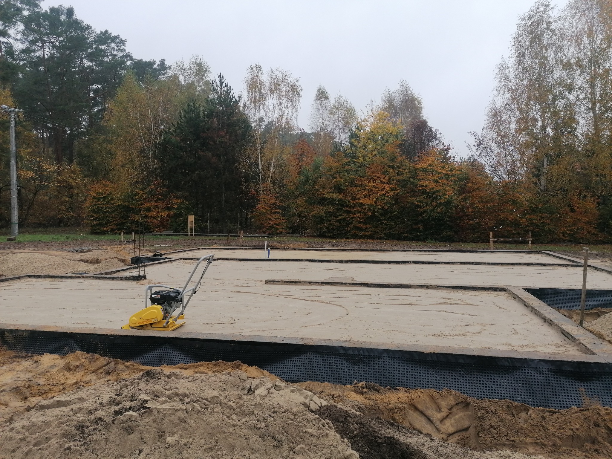 Żółta zagęszczarka wibrująca ubija piasek w przygotowanych fundamentach pod budynek, widoczne obrzeża i folia izolacyjna, w tle jesienny las.