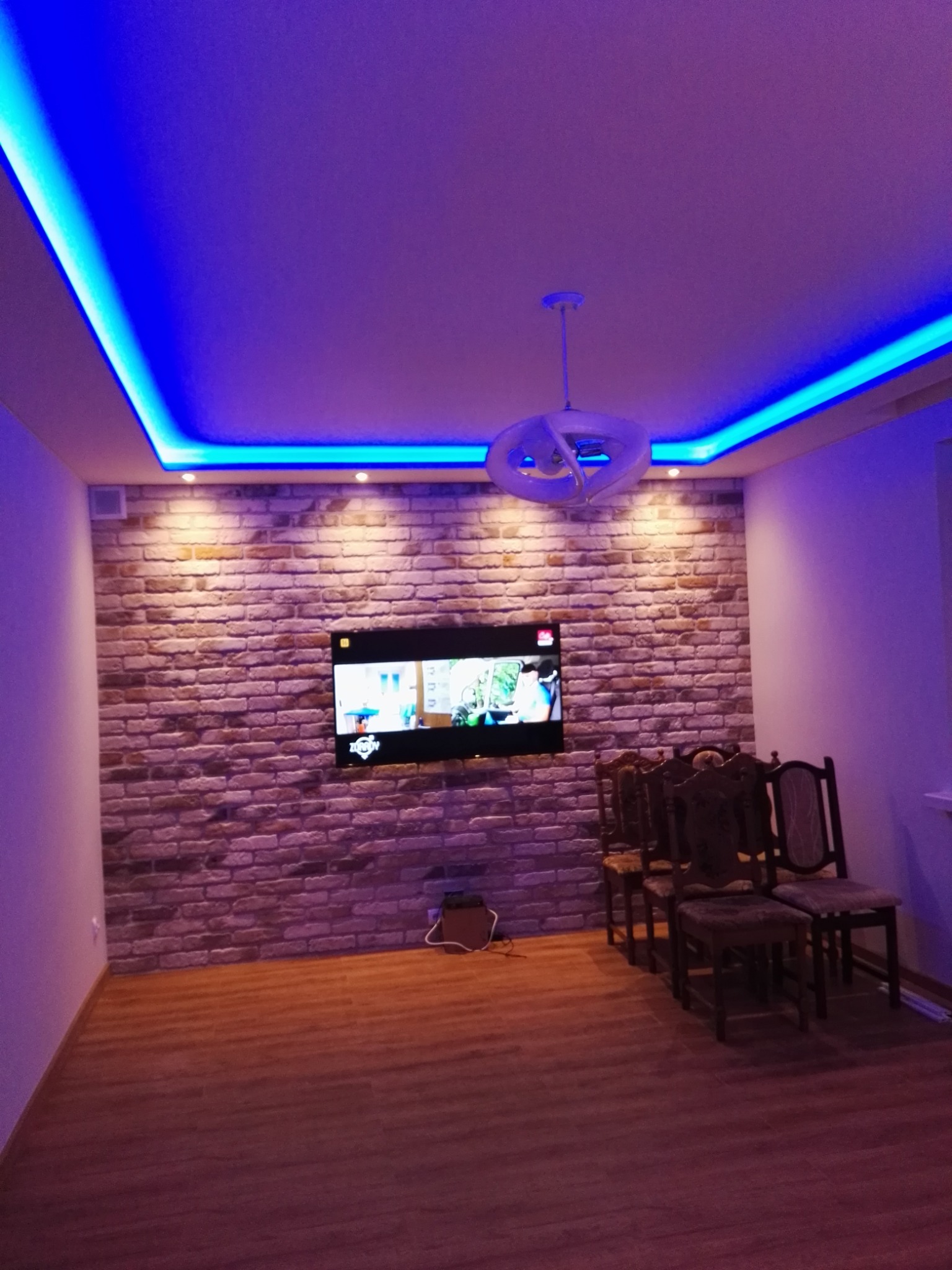 Pokój z oświetleniem LED w kolorze niebieskim, ściana wyłożona cegłą, telewizor na ścianie, drewniana podłoga i cztery krzesła.