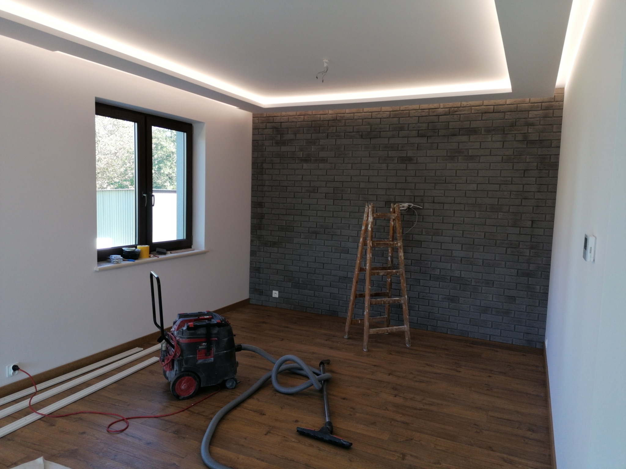Pokój po remoncie z drewnianą podłogą, ścianą z szarej cegły, oświetleniem LED na suficie, drabina drewniana oparta o ścianę, odkurzacz przemysłowy i listwy przypodłogowe przy ścianie.