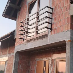 Surowa elewacja budynku w trakcie budowy z zamontowaną, minimalistyczną, ciemną balustradą okienną z płaskowników.