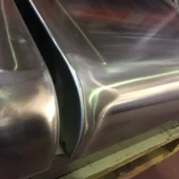 Dwa elementy metalowe, prawdopodobnie fragmenty konstrukcji, leżące na drewnianej palecie. Widoczne ślady obróbki i polerowania.