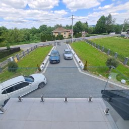 KWw - Widok z góry na posesję z dwoma samochodami na podjeździe, ogrodzeniem z elementami betonowymi i szklaną balustradą na pierwszym planie.