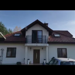 Dwupiętrowy dom z balkonem z czarną, metalową balustradą, widok z zewnątrz, częściowo zasłonięty samochodem.