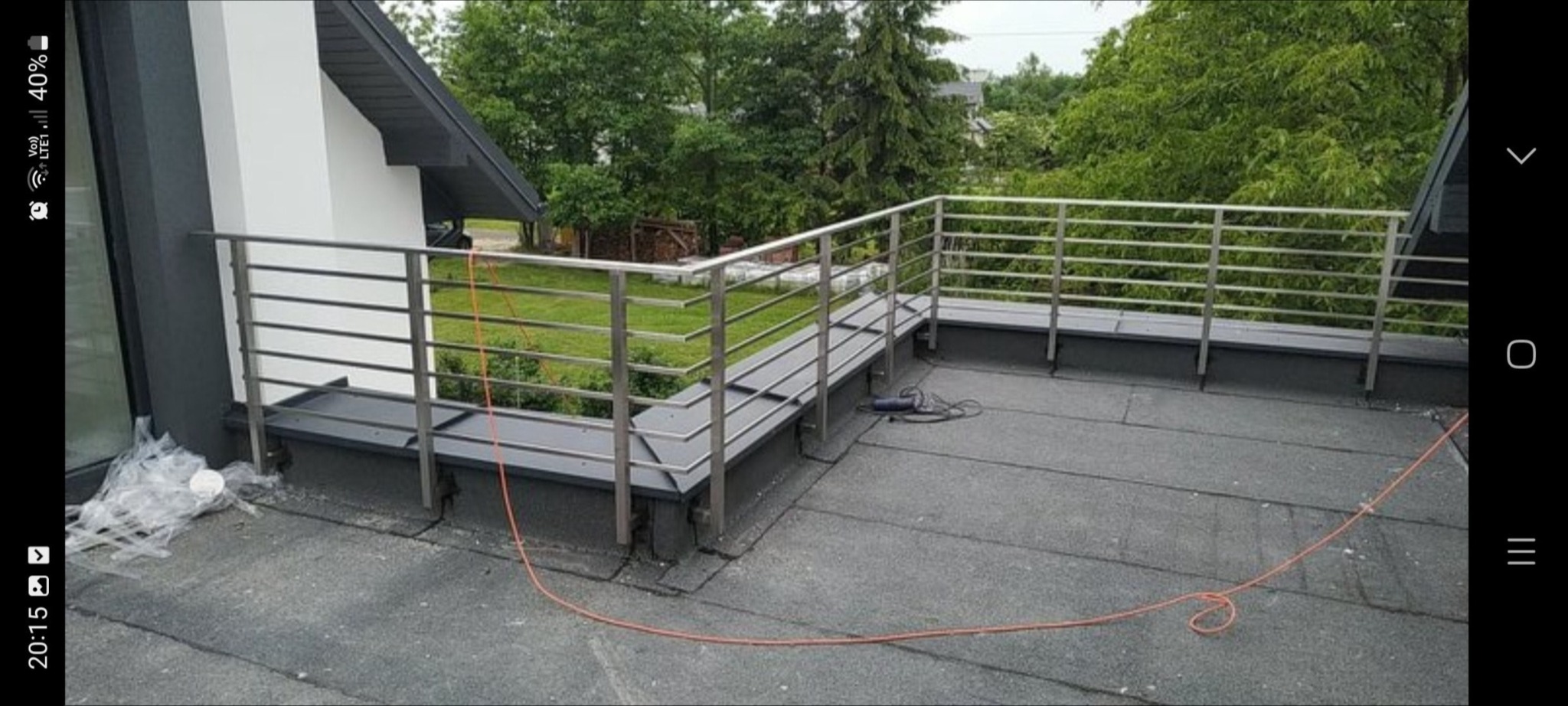 Nowoczesna balustrada ze stali na dachu budynku z widokiem na zieleń, widoczny kabel zasilający i narzędzie elektryczne.