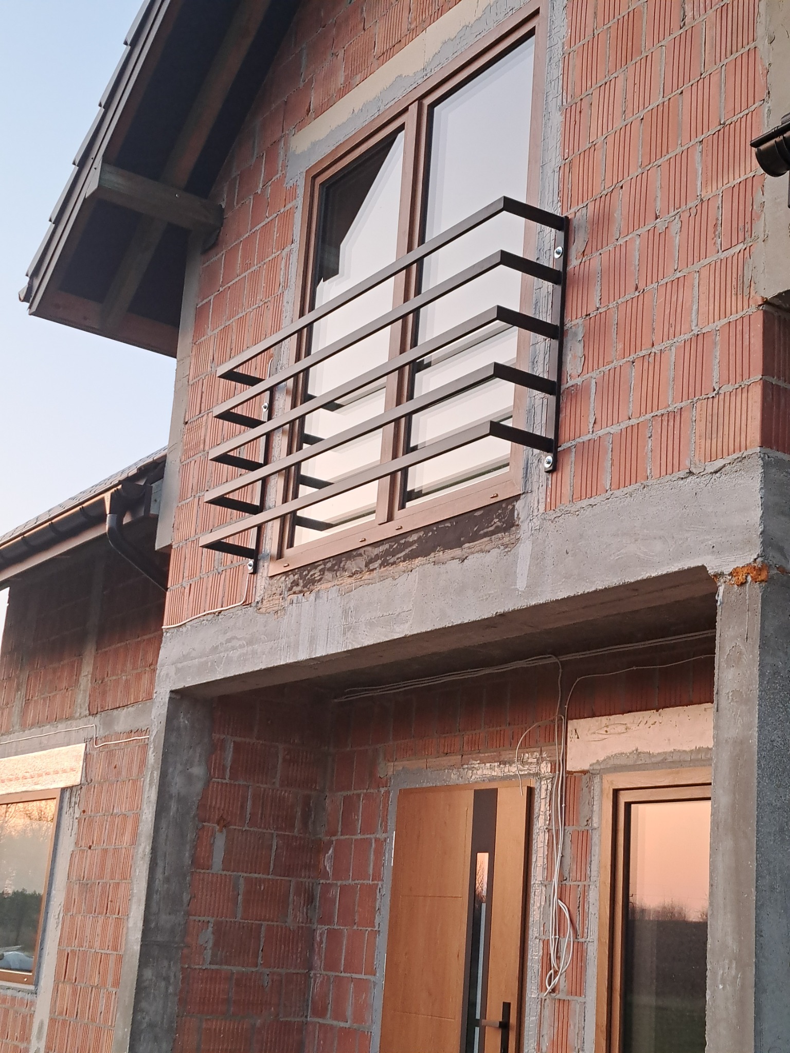 Surowa elewacja budynku w trakcie budowy z zamontowaną, minimalistyczną, ciemną balustradą okienną z płaskowników.