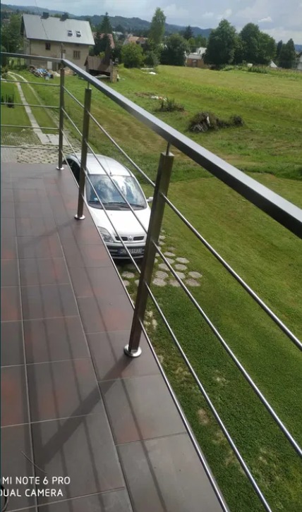 Balkon z brązowymi płytkami i nowoczesną, metalową barierką z widokiem na podjazd, samochód i zielony krajobraz