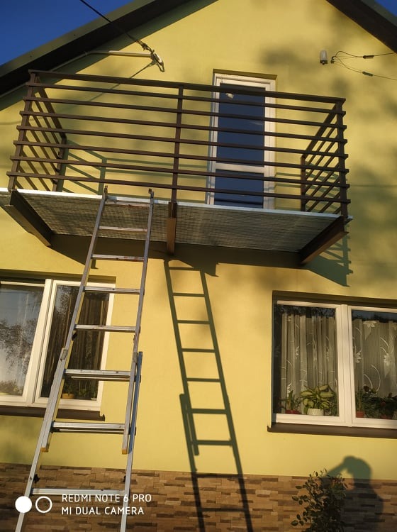 Żółta elewacja domu z zamontowanym balkonem z brązową, metalową barierką i drabina oparta o ścianę, rzucająca cień.
