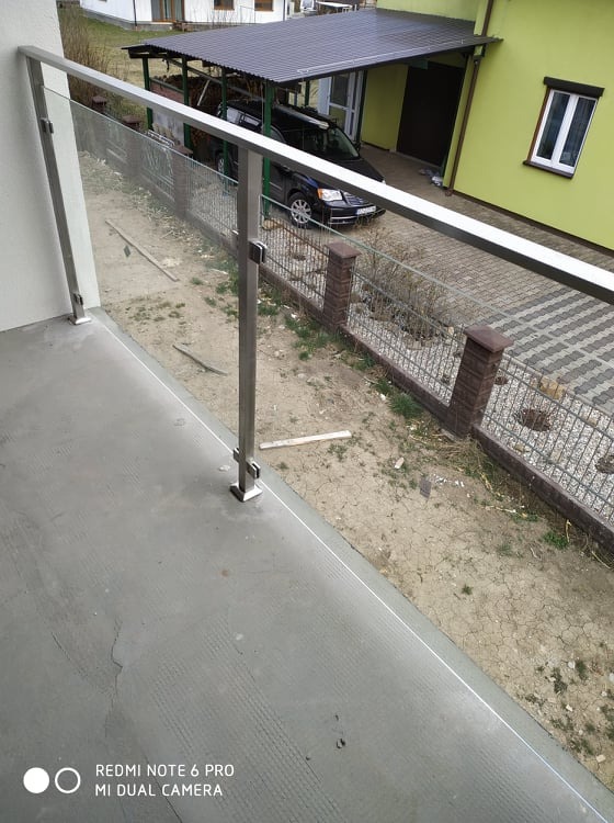 Szklana balustrada z nierdzewnymi słupkami na balkonie, widok na posesję z podjazdem i zadaszeniem dla samochodu.