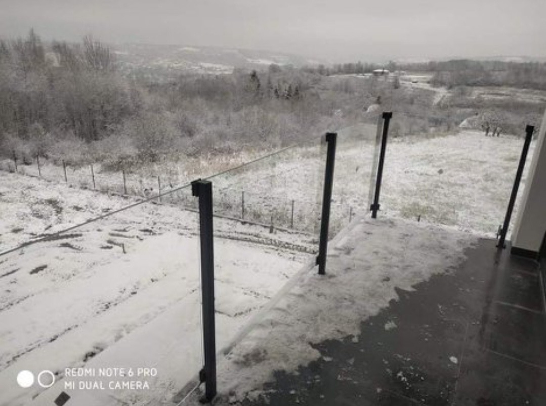 Szklana balustrada z czarnymi słupkami na tarasie pokrytym śniegiem, widok na zaśnieżony krajobraz w tle.