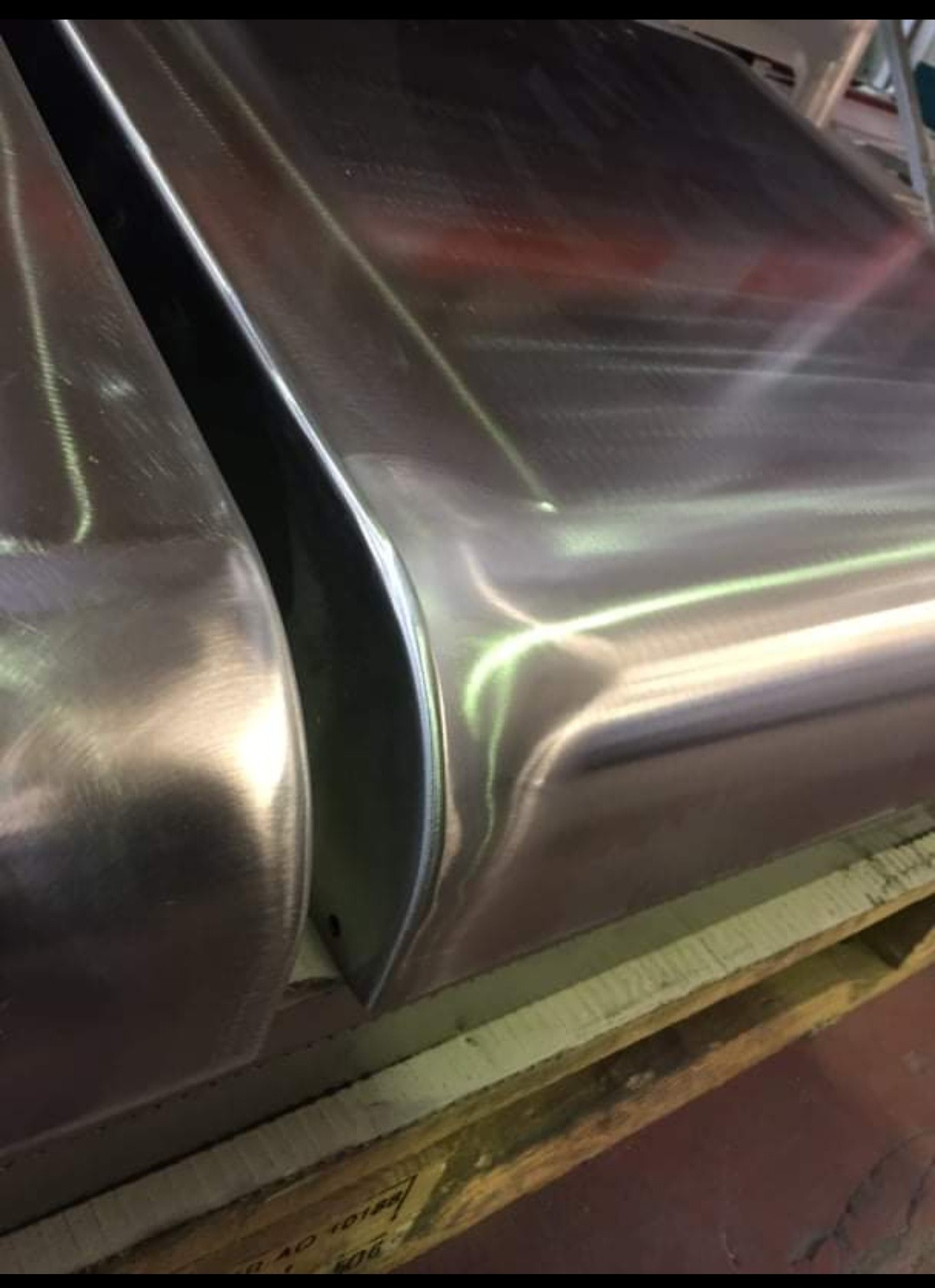 Dwa elementy metalowe, prawdopodobnie fragmenty konstrukcji, leżące na drewnianej palecie. Widoczne ślady obróbki i polerowania.