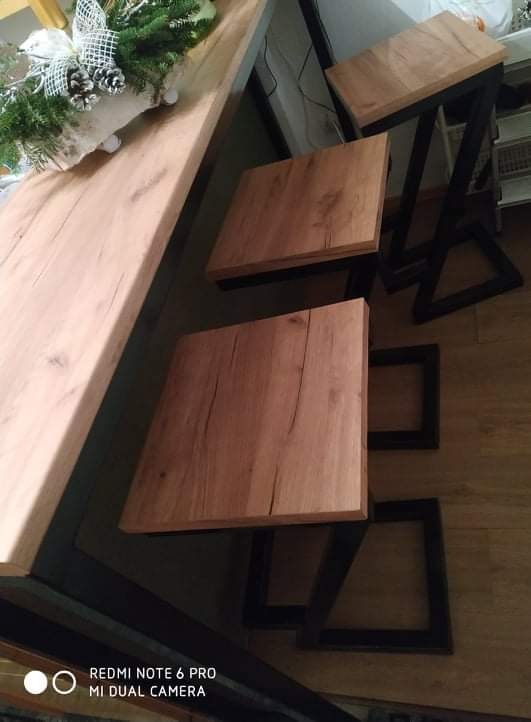 Stół barowy z blatem o fakturze drewna i metalowymi nogami oraz dwa drewniane stołki barowe z czarnymi metalowymi podstawami o geometrycznym kształcie, uzupełnione świąteczną dekoracją.