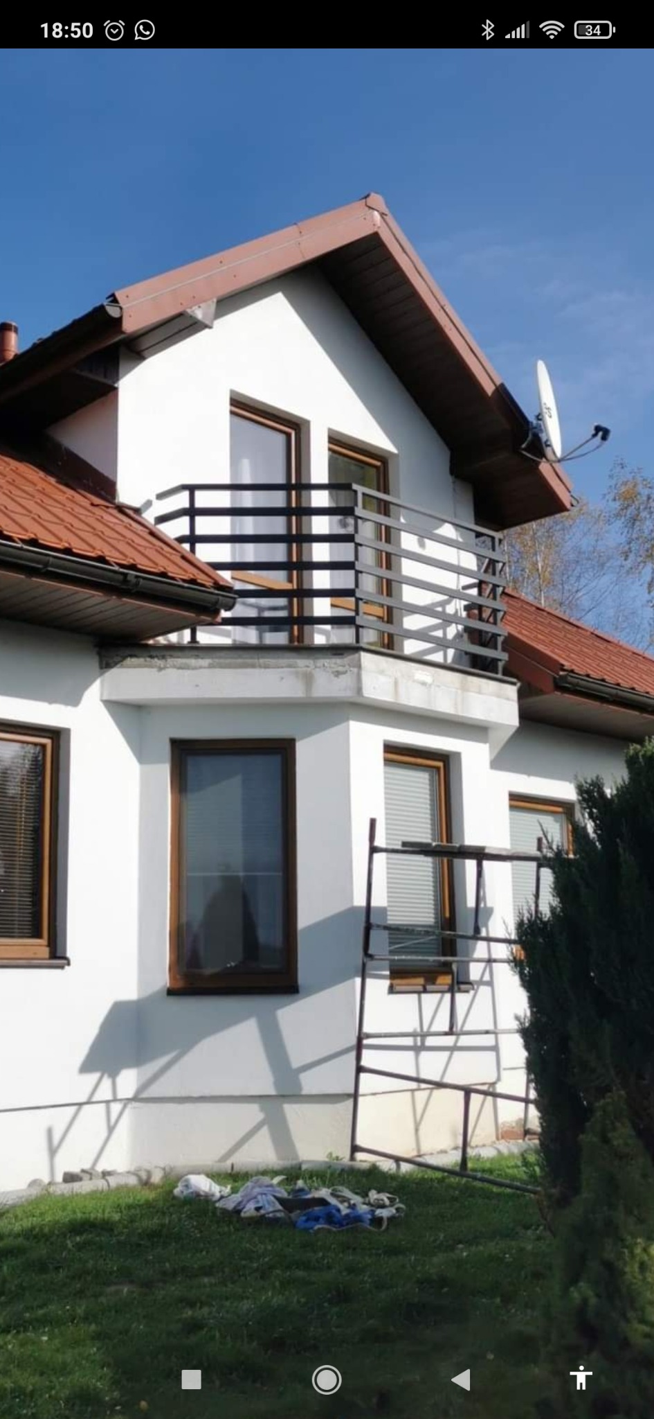 Czarna balustrada balkonowa z płaskowników zamontowana w Rzeszowie przy białym domu z brązowymi oknami i czerwoną dachówką, widok z dołu na balkon z metalową konstrukcją i anteną satelitarną.