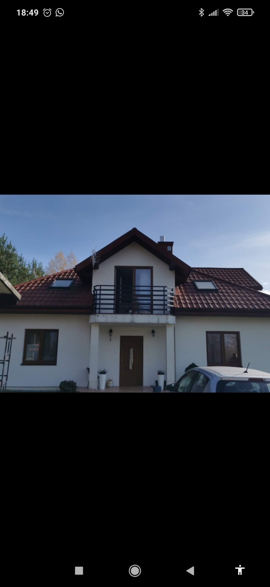 Dwupiętrowy dom z balkonem z czarną, metalową balustradą, widok z zewnątrz, częściowo zasłonięty samochodem.