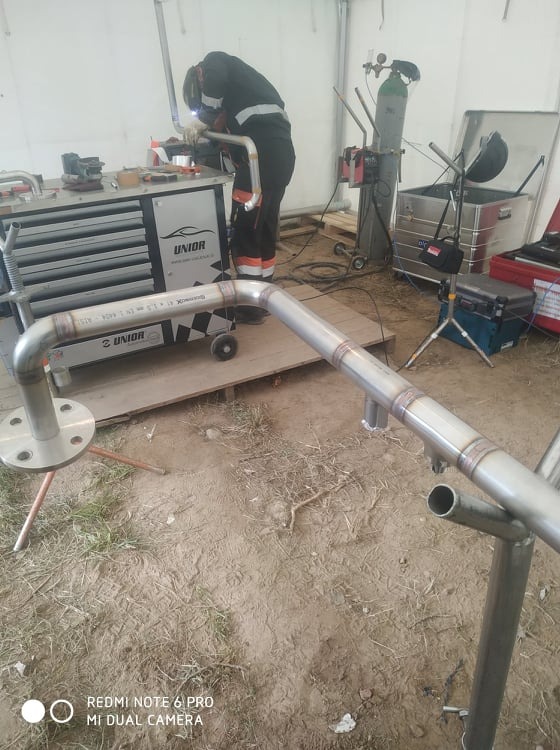 Pracownik w kombinezonie spawalniczym łączy elementy rur ze stali nierdzewnej metodą spawania, widoczny sprzęt spawalniczy, w tle wózek narzędziowy marki Unior i butla z gazem technicznym.