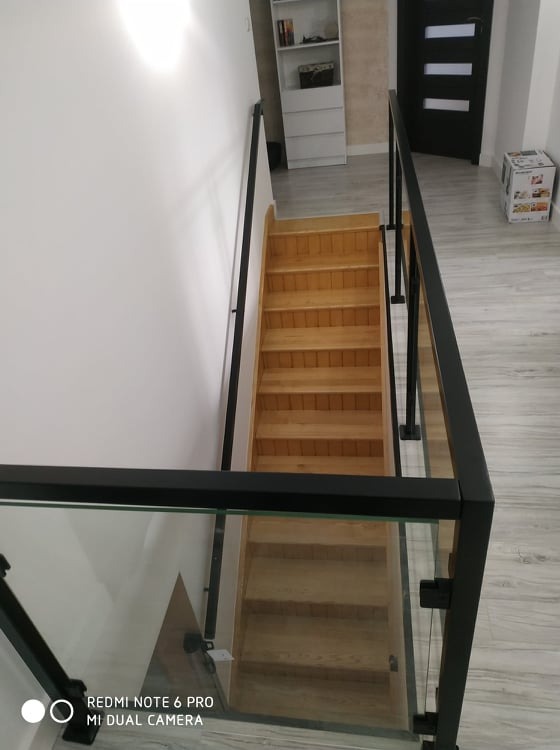 Drewniane schody z czarną, metalową balustradą i szklanym wypełnieniem, widok z góry na dół, minimalistyczny design, jasne wnętrze.