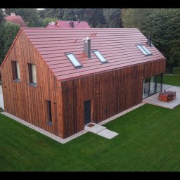 BLACKWOOD - Nowoczesny dom z elewacją z ciemnego, opalanego drewna (Shou Sugi Ban), czerwonym dachem i tarasem z jasnego drewna, otoczony zielonym trawnikiem. Widok z góry.