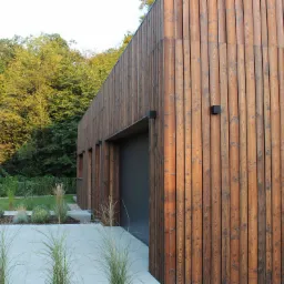 Nowoczesny dom z elewacją z pionowych desek w odcieniach brązu, z szarą bramą garażową i minimalistycznym ogrodem z ozdobnymi trawami.