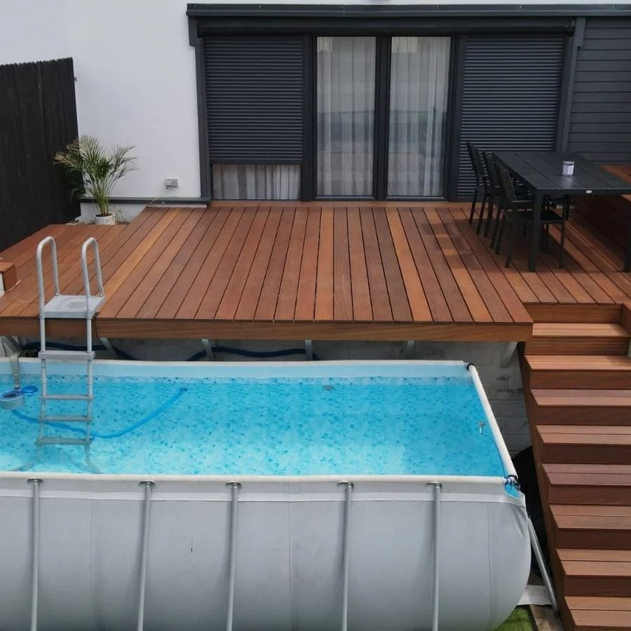 Taras z desek egzotycznych otaczający basen naziemny, z widocznymi schodami i meblami ogrodowymi na tle nowoczesnego domu z roletami zewnętrznymi.