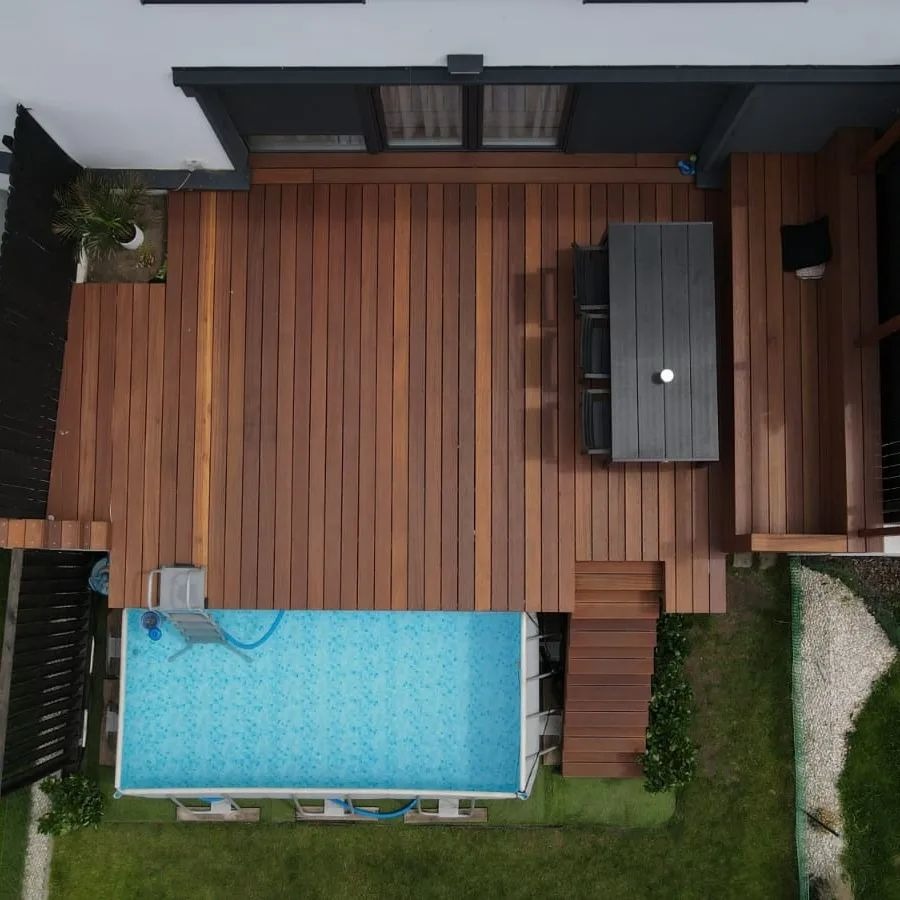 Widok z góry na taras z desek w odcieniach brązu, przylegający do domu z basenem; na tarasie stół z krzesłami, obok schody.