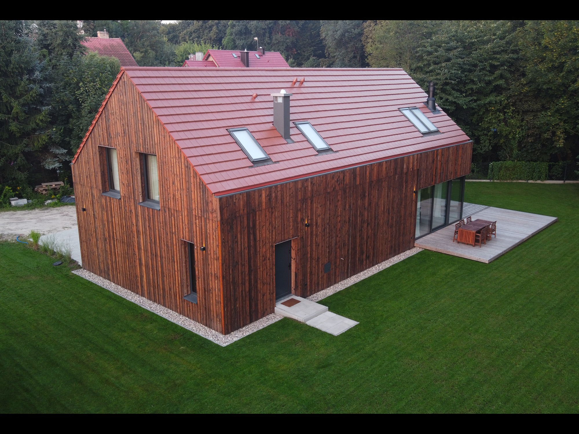 Nowoczesny dom z elewacją z ciemnego, opalanego drewna (Shou Sugi Ban), czerwonym dachem i tarasem z jasnego drewna, otoczony zielonym trawnikiem. Widok z góry.