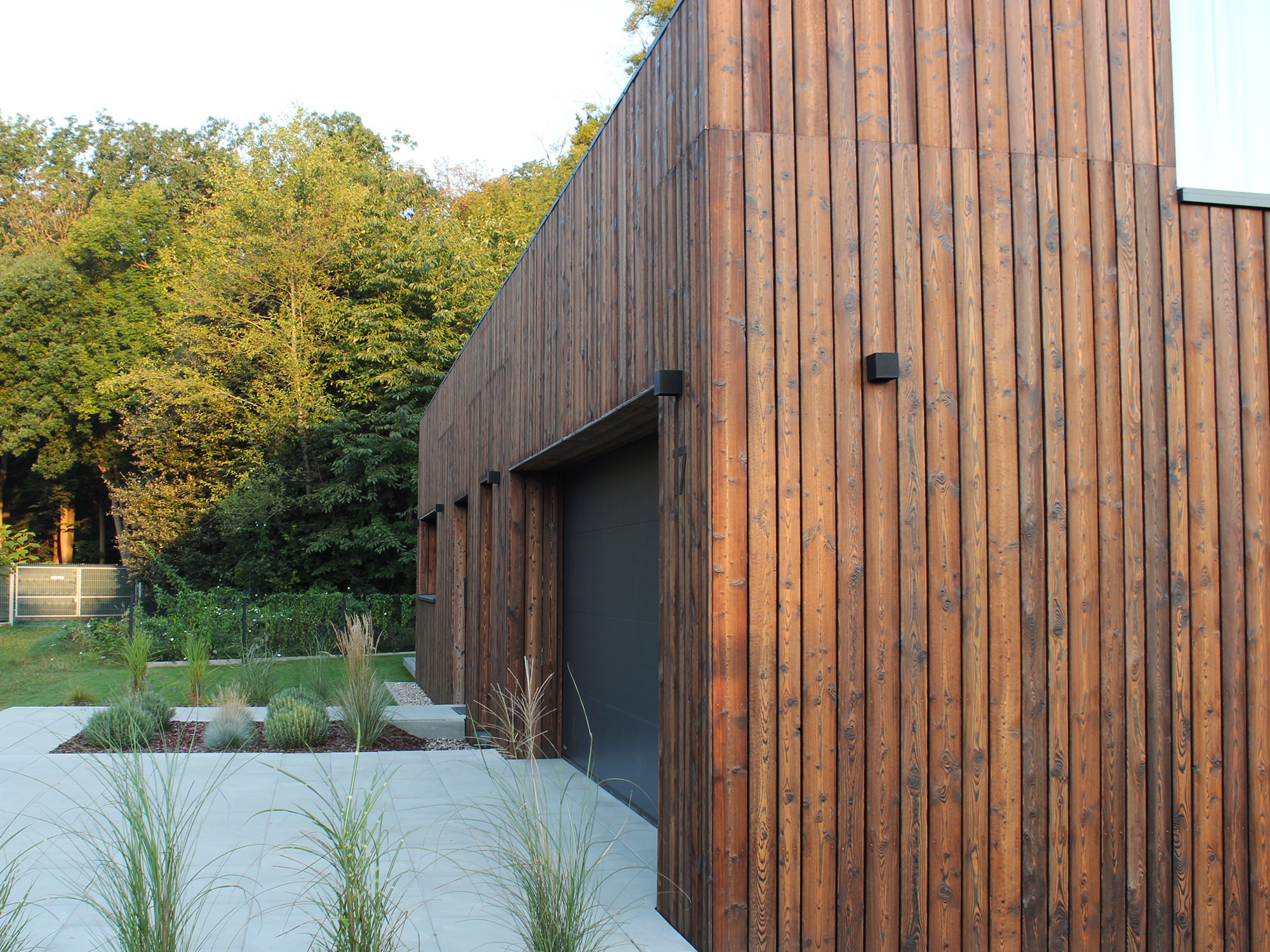 Nowoczesny dom z elewacją z pionowych desek w odcieniach brązu, z szarą bramą garażową i minimalistycznym ogrodem z ozdobnymi trawami.