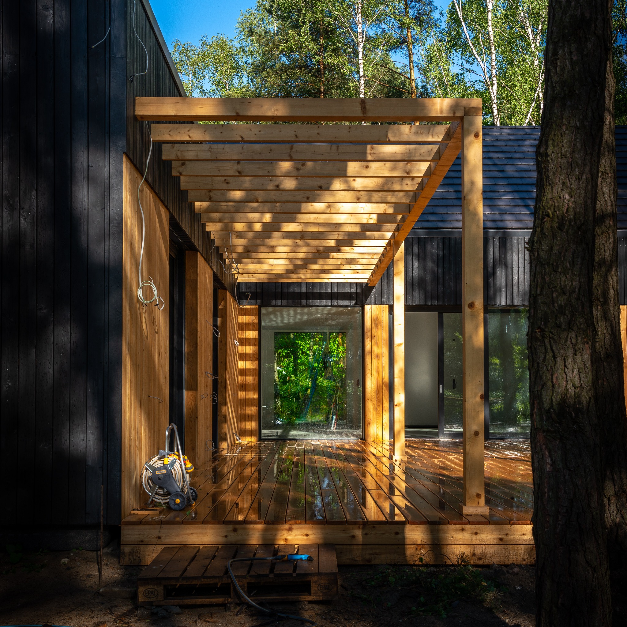 Nowoczesny dom z ciemną elewacją i jasną, drewnianą pergolą oraz tarasem po deszczu, na którym odbijają się promienie słoneczne, widoczna myjka ciśnieniowa.