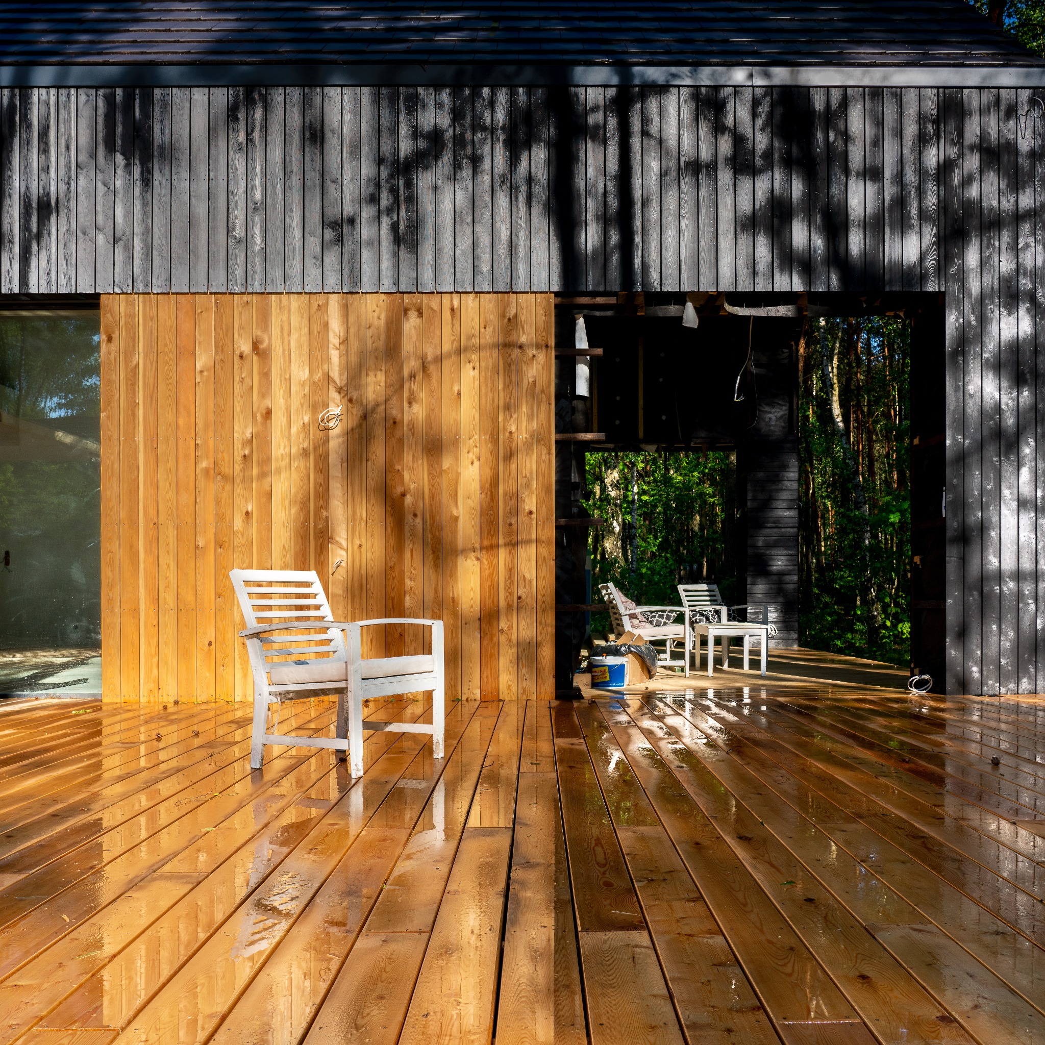 Nowoczesny taras drewniany z jasnego drewna po deszczu, białe krzesło na pierwszym planie, w tle widoczny fragment elewacji z ciemnego drewna i zieleń lasu