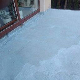 Jurex - Szara, świeżo nałożona warstwa hydroizolacyjna na balkonie, widoczna przy brązowych ramach okiennych, z rośliną doniczkową w tle za szybą.