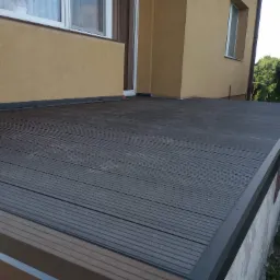 Szara deska kompozytowa tworząca taras przylegający do budynku z oknami, widoczne detale łączeń i wykończenia krawędzi.