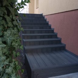 Jurex - Schody zewnętrzne wyłożone deską kompozytową w kolorze grafitowym, prowadzące do budynku z elewacją w odcieniach beżu i różu, częściowo zasłonięte zielonym bluszczem.