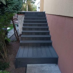 Jurex - Schody zewnętrzne z kompozytu drewna, prowadzące do budynku o elewacji w odcieniach beżu i różu, otoczone zielenią.