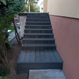 Schody zewnętrzne z kompozytu drewna, prowadzące do budynku o elewacji w odcieniach beżu i różu, otoczone zielenią.