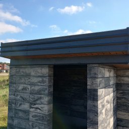 Jurex - Gotowa konstrukcja z szarych, wzorzystych betonowych bloczków z metalowym dachem na tle błękitnego nieba i krajobrazu z domem w oddali.