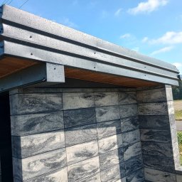 Jurex - Zadaszona konstrukcja z szarych, wzorzystych bloczków betonowych i drewnianym podbitką, widoczna na tle błękitnego nieba i pola.