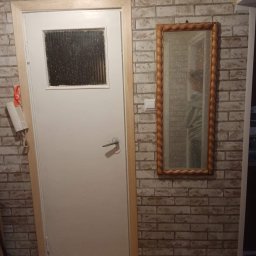 Jurex - Ściana wykończona ceglastymi płytkami imitującymi mur, obok drzwi z okienkiem wisi lustro w ozdobnej ramie.