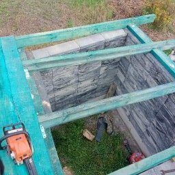 Jurex - Wykop wyłożony płytami imitującymi kamień, z drewnianą konstrukcją nad nim, widoczna piła łańcuchowa i narzędzia wewnątrz.