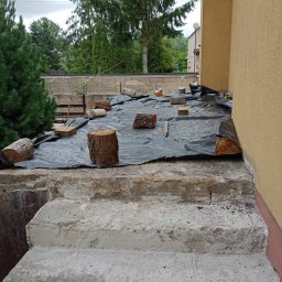 Jurex - Przygotowanie do izolacji: betonowe schody zabezpieczone czarną folią, obciążoną kawałkami drewna, widok z góry na tle zieleni i elewacji budynku.
