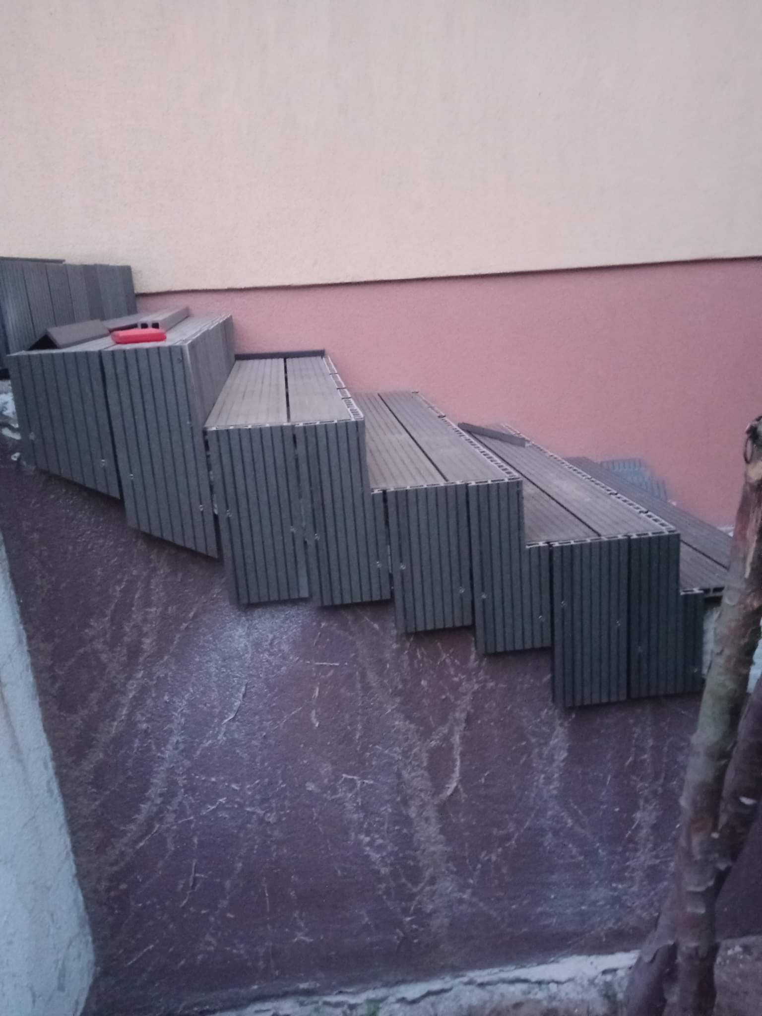 Stopnie schodów wykonane z kompozytowych desek tarasowych, ułożone na nierównej powierzchni z widocznym poziomowaniem i czerwoną poziomnicą na górnym stopniu, tło stanowi ściana budynku w dwóch...