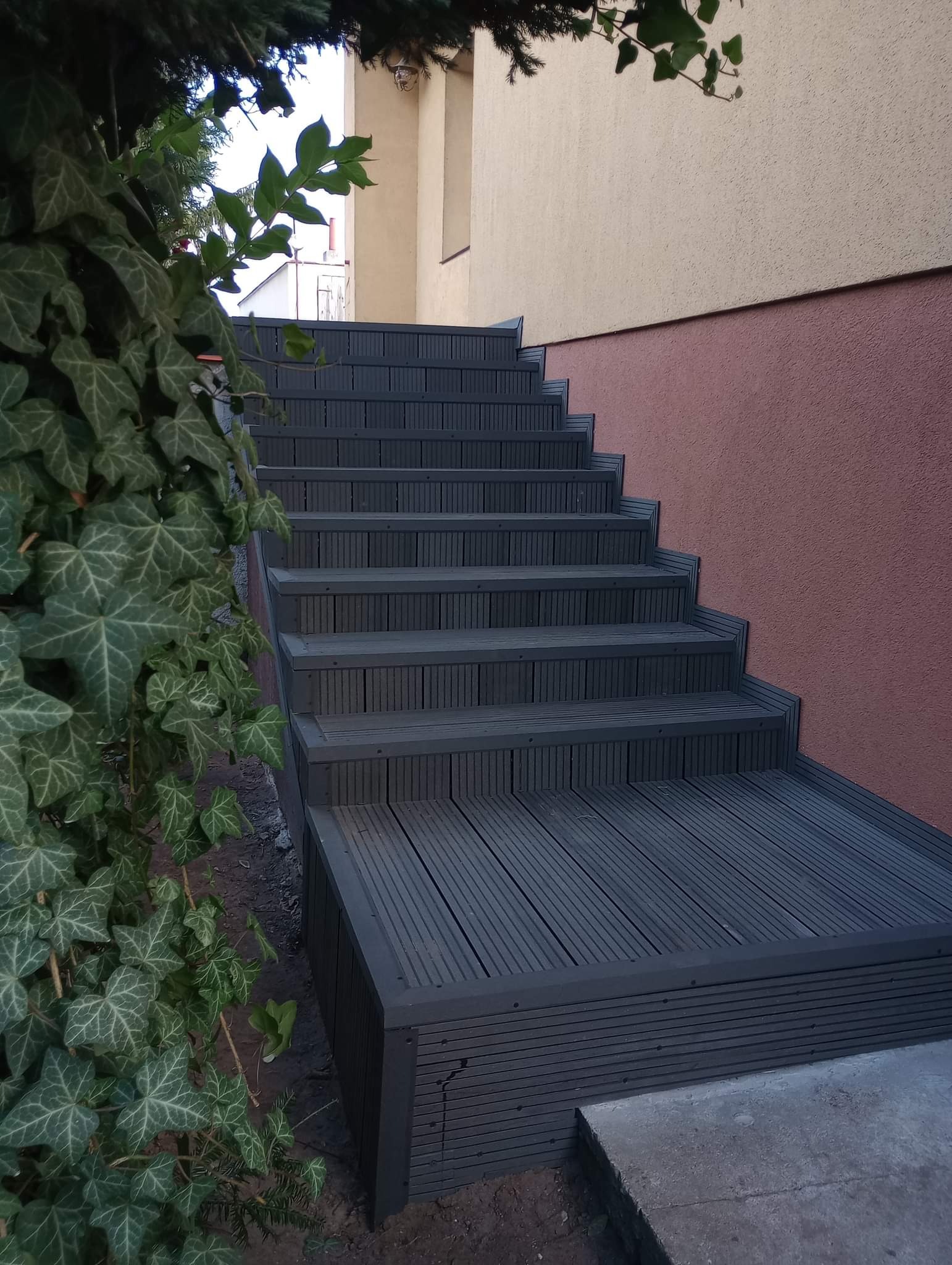 Schody zewnętrzne wyłożone deską kompozytową w kolorze grafitowym, prowadzące do budynku z elewacją w odcieniach beżu i różu, częściowo zasłonięte zielonym bluszczem.