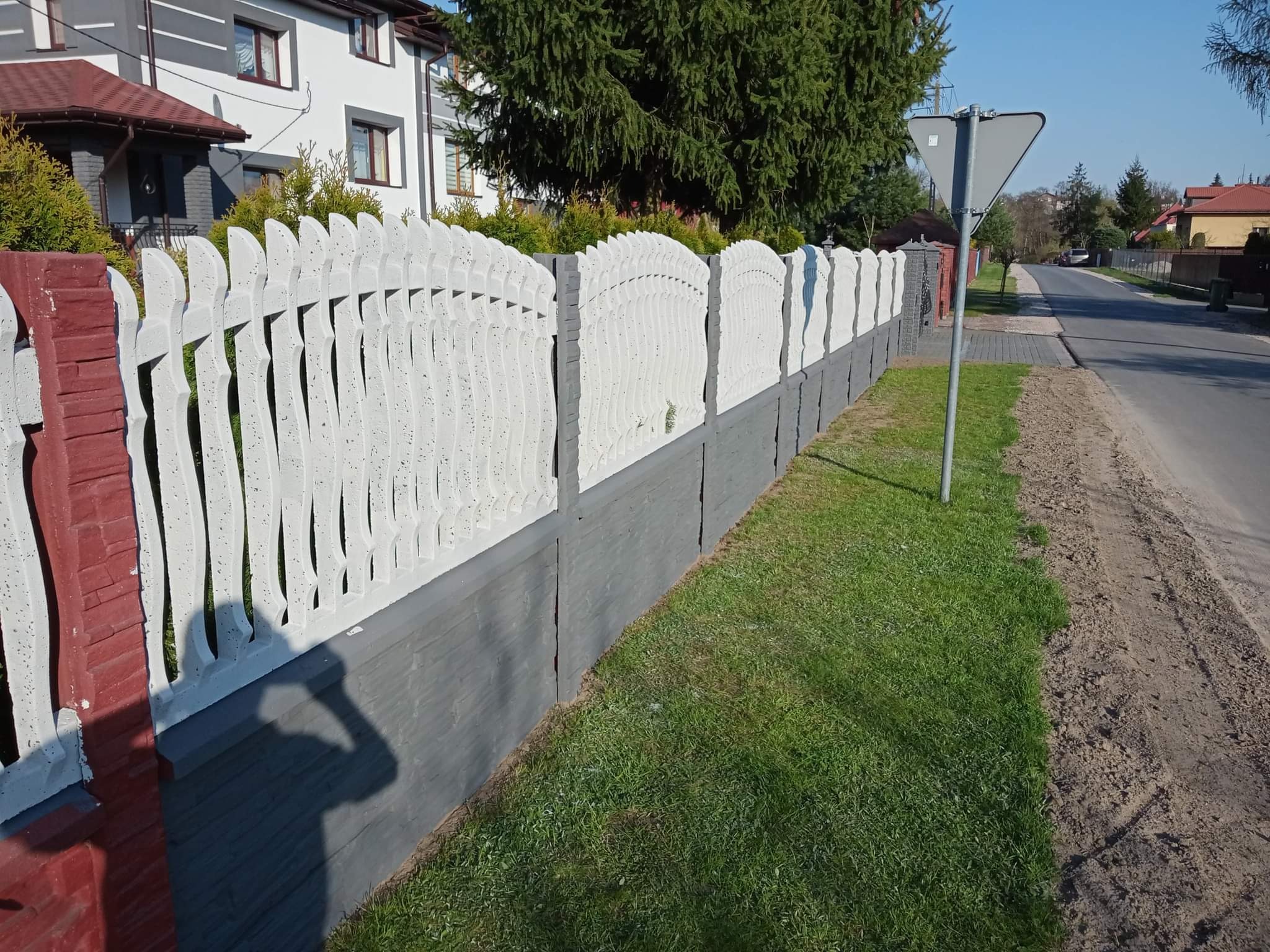 Nowoczesne ogrodzenie betonowe z dekoracyjnymi przęsłami w kolorze białym i szarą podmurówką, zakończone ceglanym słupkiem, oddzielające posesję od ulicy.