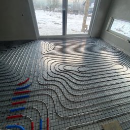 HYDRO-MAX Marcin Adamczyk - Ułożone rury ogrzewania podłogowego na siatce montażowej w jasnym pomieszczeniu z dużym oknem, za którym widać zimowy krajobraz. Fragmenty rur w kolorze niebieskim i czerwonym.