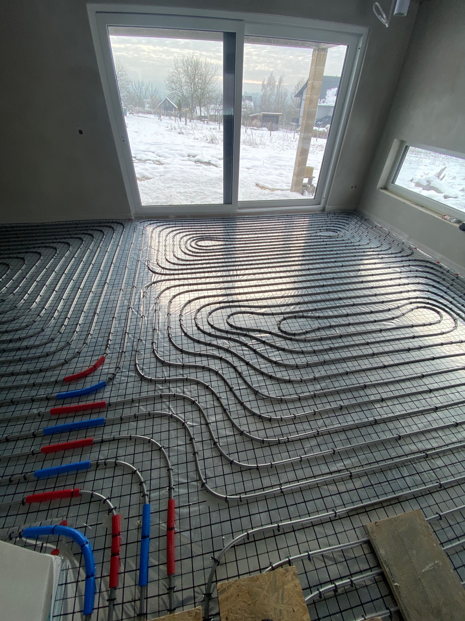 Ułożone rury ogrzewania podłogowego na siatce montażowej w jasnym pomieszczeniu z dużym oknem, za którym widać zimowy krajobraz. Fragmenty rur w kolorze niebieskim i czerwonym.