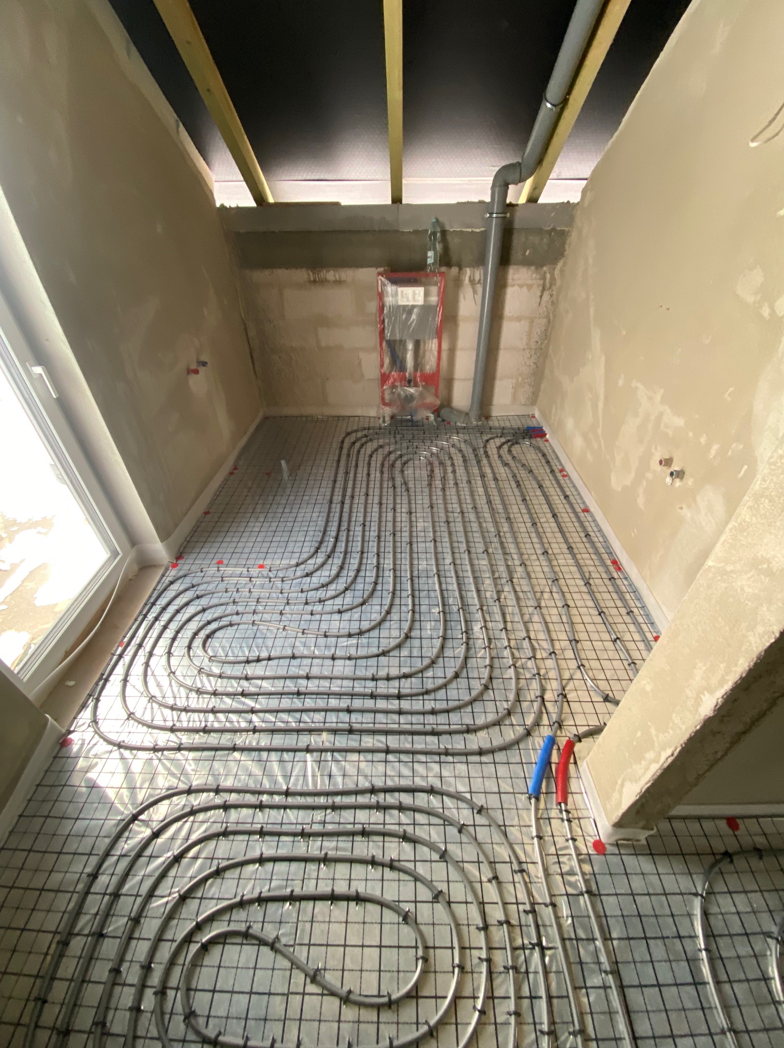 Szare rury ogrzewania podłogowego ułożone na czarnej siatce w surowym wnętrzu z oknem. Widoczne elementy instalacji i sufit z drewnianymi belkami.