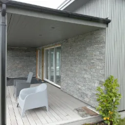 Zadaszony taras z elewacją z kamienia dekoracyjnego, jasna drewniana podłoga, szare krzesła ogrodowe, widoczne przesuwne drzwi tarasowe i fragment drewnianej ściany budynku.
