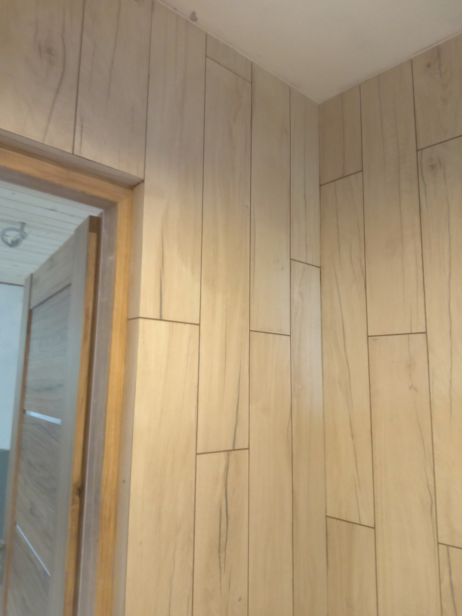 Ściana wyłożona panelami imitującymi drewno, z widoczną częścią ościeżnicy i fragmentem drzwi w tle.